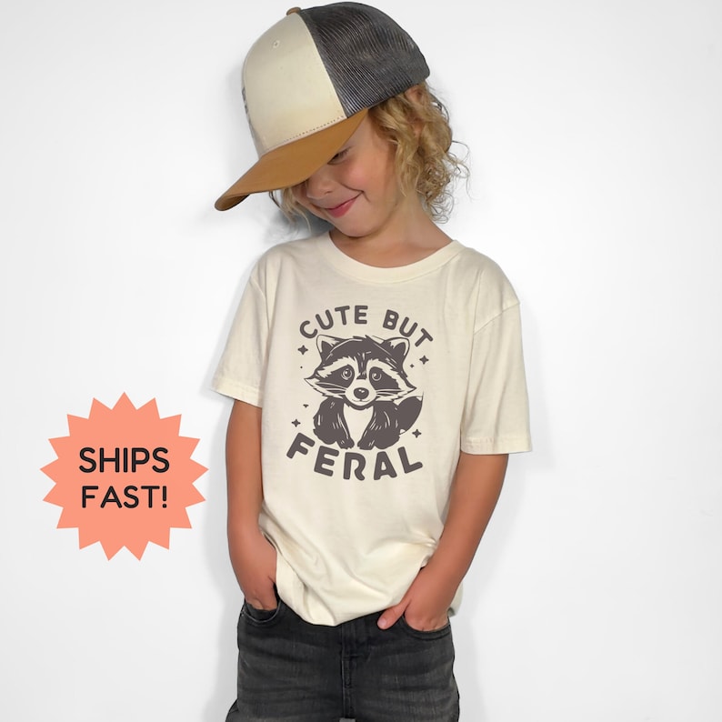K&ouml;nnte beinhalten: Ein Kind tr&auml;gt ein cremefarbenes T-Shirt mit einer Waschb&auml;r-Grafik und dem Text "Cute But Feral". Das Kind tr&auml;gt auch eine Trucker-M&uuml;tze mit braunem Schirm und grauem Mesh-R&uuml;cken. Das Kind tr&auml;gt dunkelgraue Jeans.