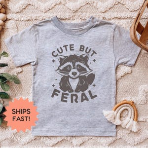 K&ouml;nnte beinhalten: Ein hellgraues T-Shirt mit einem Waschb&auml;r-Motiv und dem Text "CUTE BUT FERAL". Der Waschb&auml;r ist in Graut&ouml;nen dargestellt. Das T-Shirt hat kurze &Auml;rmel und einen Rundhalsausschnitt. Das T-Shirt wird auf einer beigen Oberfl&auml;che pr&auml;sentiert.