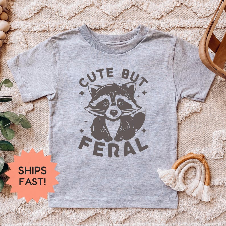 K&ouml;nnte beinhalten: Ein hellgraues T-Shirt mit einem Waschb&auml;r-Motiv und dem Text "CUTE BUT FERAL". Der Waschb&auml;r ist in Graut&ouml;nen dargestellt. Das T-Shirt hat kurze &Auml;rmel und einen Rundhalsausschnitt. Das T-Shirt wird auf einer beigen Oberfl&auml;che pr&auml;sentiert.