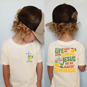 May include: A child wearing a cream-colored t-shirt and a brown and tan trucker hat. The front of the shirt has a blue cross, a lemon, and the name "Clark". The back of the shirt says "Life gave me some lemons but my Jesus he be makin' lemonade."