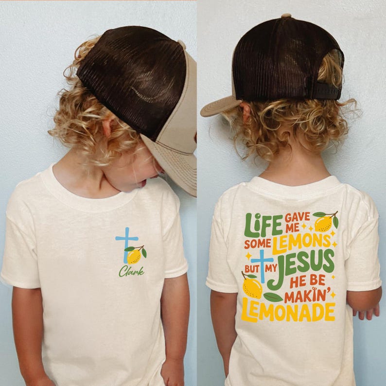 May include: A child wearing a cream-colored t-shirt and a brown and tan trucker hat. The front of the shirt has a blue cross, a lemon, and the name "Clark". The back of the shirt says "Life gave me some lemons but my Jesus he be makin' lemonade."