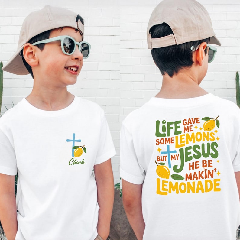 May include: White t-shirt with a cross and lemon graphic. The front of the shirt has a blue cross and the name "Clark" in green script. The back of the shirt has the text "Life Gave Me Some Lemons But My Jesus He Be Makin' Lemonade."
