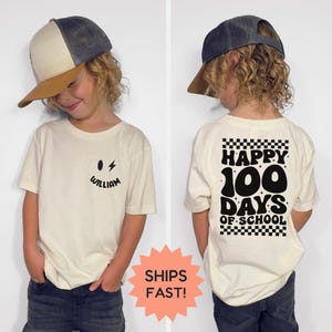 May include: Cream-coloured t-shirt featuring a black smiley face, lightning bolt, and the name "William" on the front. The back of the shirt displays "Happy 100 Days of School" in a retro font. The shirt is worn by a child.