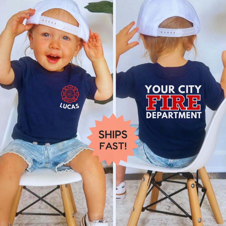 May include: A navy blue t-shirt with a red fire department emblem and the name "Lucas" printed on the front. The back of the shirt displays "YOUR CITY FIRE DEPARTMENT" in white. The child is wearing a white baseball cap and denim shorts.