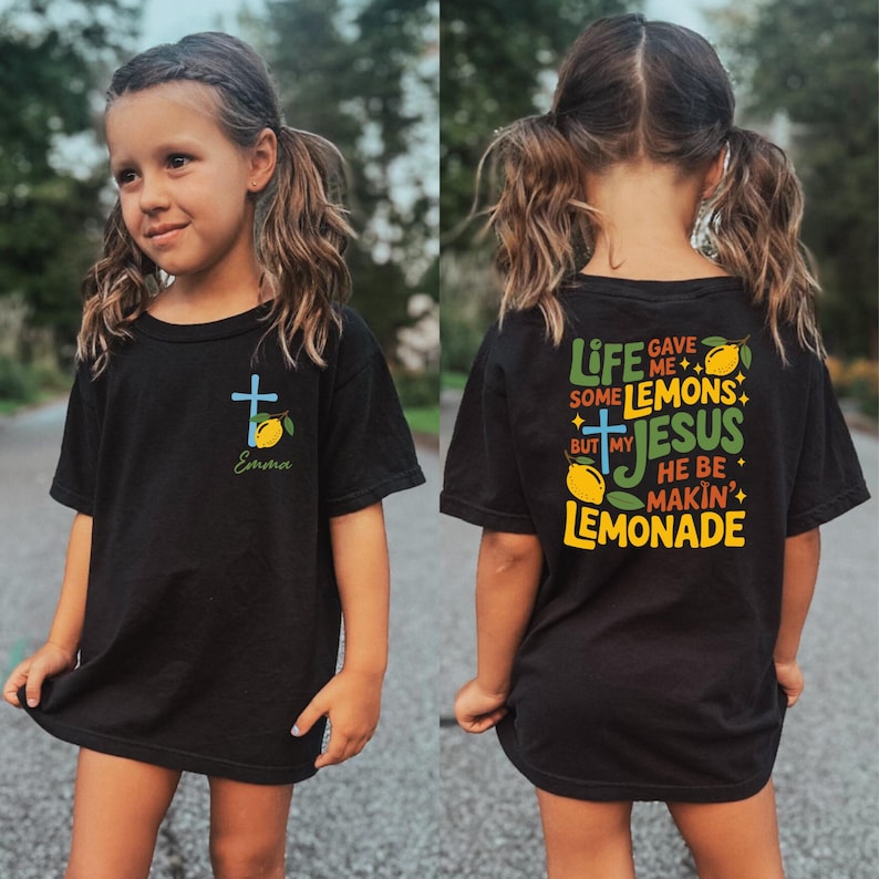 May include: A black t-shirt with a cross and lemon graphic, and the name "Emma" in script. The back of the shirt has the text "Life gave me some lemons but my Jesus he be makin' lemonade" with lemon and cross graphics.