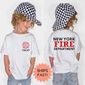 May include: White t-shirt with a red and black New York Fire Department logo and the name "LUCAS" printed on the front. The back of the shirt displays "NEW YORK FIRE DEPARTMENT" in black and red. The child is wearing a black and white checkered baseball cap.