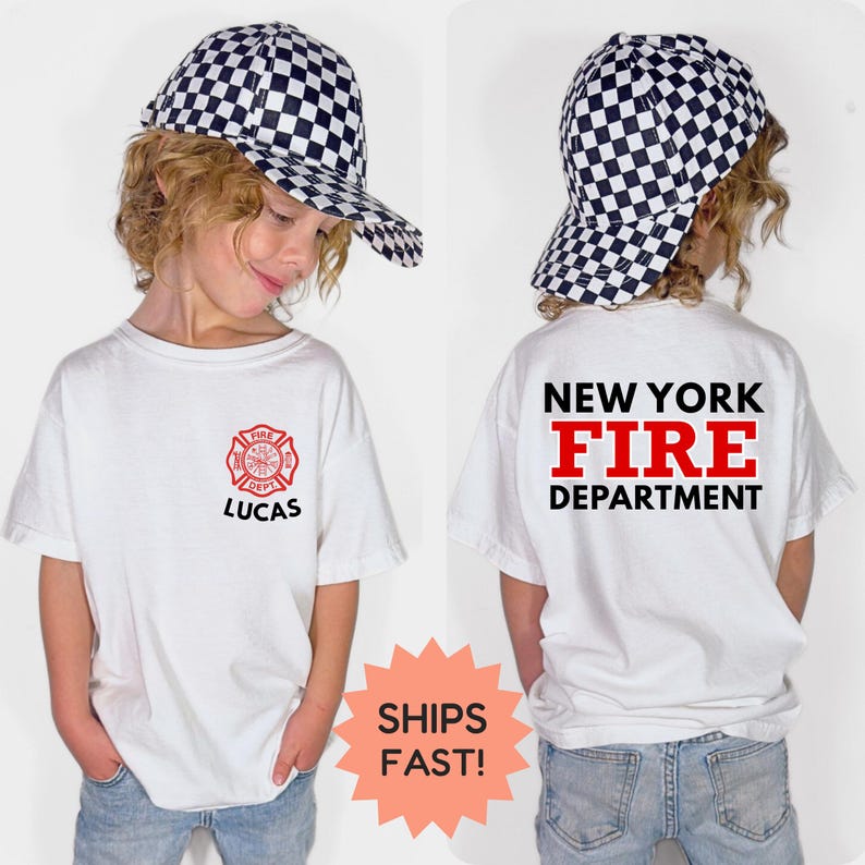 May include: White t-shirt with a red and black New York Fire Department logo and the name "LUCAS" printed on the front. The back of the shirt displays "NEW YORK FIRE DEPARTMENT" in black and red. The child is wearing a black and white checkered baseball cap.