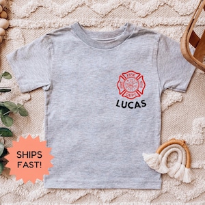 May include: A light gray t-shirt featuring a red and black firefighter emblem with the text "FIRE DEPT" and the name "LUCAS" in black. The shirt is short-sleeved and has a crew neck.