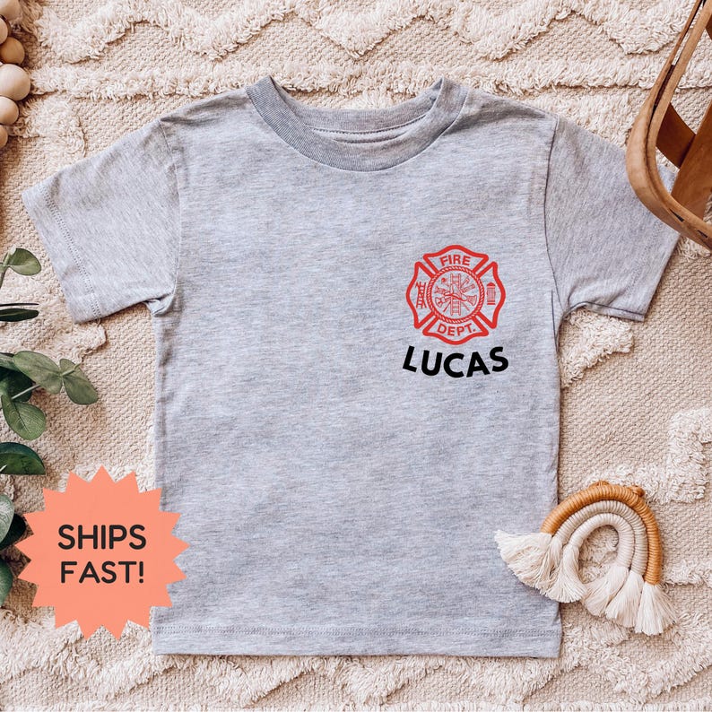 May include: A light gray t-shirt featuring a red and black firefighter emblem with the text "FIRE DEPT" and the name "LUCAS" in black. The shirt is short-sleeved and has a crew neck.
