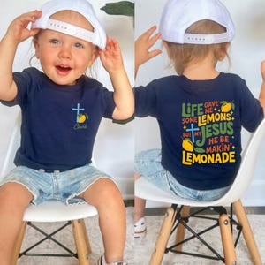 May include: Navy blue t-shirt with a cross and lemon graphic, plus the name "Clark". The back of the shirt says "Life gave me some lemons but my Jesus he be makin' lemonade". The child is wearing a white baseball cap and denim shorts.