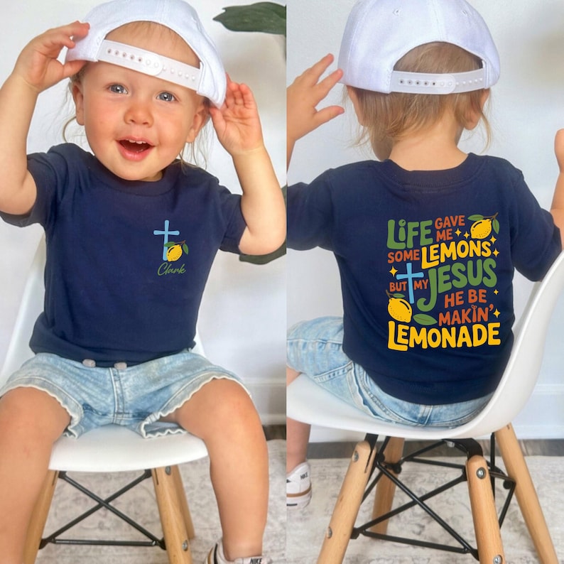 May include: Navy blue t-shirt with a cross and lemon graphic, plus the name "Clark". The back of the shirt says "Life gave me some lemons but my Jesus he be makin' lemonade". The child is wearing a white baseball cap and denim shorts.