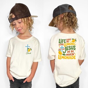 May include: Off-white short-sleeve t-shirt with a blue cross and lemon graphic. The front of the shirt has the name "Clark" below the graphic. The back of the shirt has the text "Life gave me some lemons but my Jesus he be makin' lemonade."