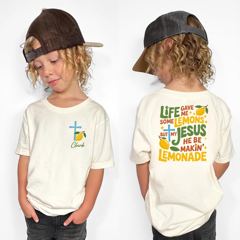 May include: Off-white short-sleeve t-shirt with a blue cross and lemon graphic. The front of the shirt has the name "Clark" below the graphic. The back of the shirt has the text "Life gave me some lemons but my Jesus he be makin' lemonade."