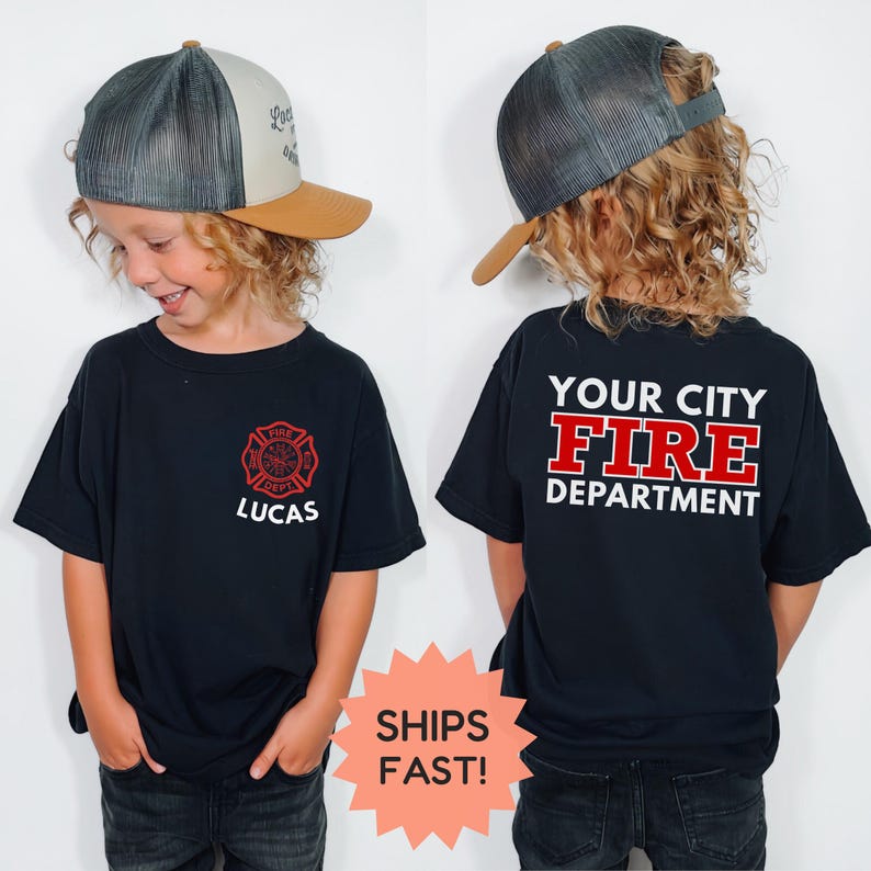 May include: Black t-shirt with a red and white fire department emblem and the name "Lucas". The back of the shirt reads "Your City Fire Department" in white and red lettering. The child is wearing a gray and tan trucker hat.
