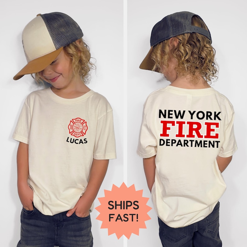 May include: A cream-colored t-shirt with a red fire department emblem and the name "LUCAS" printed below it. The back of the shirt displays "NEW YORK FIRE DEPARTMENT" in black and red. The child is wearing a trucker hat.