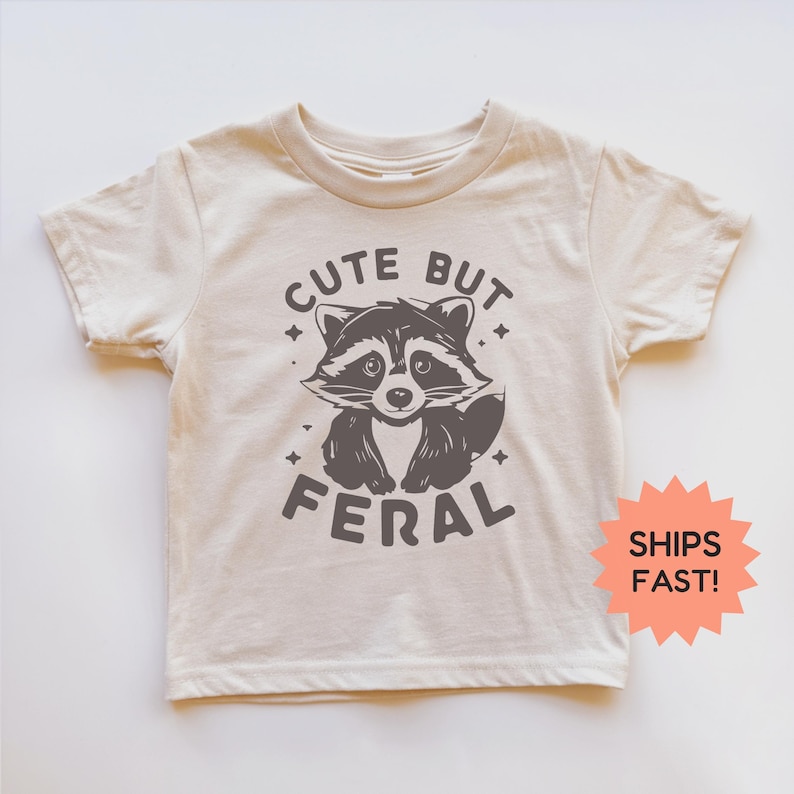 K&ouml;nnte beinhalten: Cremefarbenes Kinder-T-Shirt mit einem grauen Waschb&auml;r-Motiv und dem Schriftzug "Cute But Feral". Das Design beinhaltet Sterne. Das T-Shirt hat kurze &Auml;rmel und einen Rundhalsausschnitt.