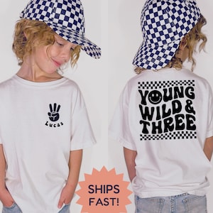 May include: White t-shirt with a black peace sign and the name "Lucas" printed on the front. The back of the shirt features the text "Young Wild & Three" with a smiley face and checkered borders. The child is wearing a checkered hat.