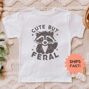 K&ouml;nnte beinhalten: Wei&szlig;es T-Shirt mit einer grauen Waschb&auml;r-Grafik und dem Text "CUTE BUT FERAL". Die Waschb&auml;r-Illustration hat ein verspieltes Design. Das T-Shirt ist aus einem weichen Material.