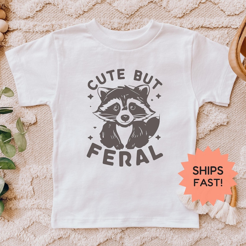 K&ouml;nnte beinhalten: Wei&szlig;es T-Shirt mit einer grauen Waschb&auml;r-Grafik und dem Text "CUTE BUT FERAL". Die Waschb&auml;r-Illustration hat ein verspieltes Design. Das T-Shirt ist aus einem weichen Material.