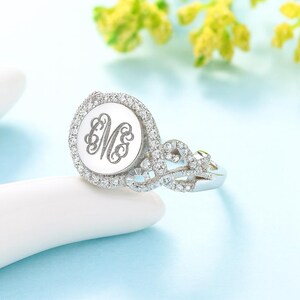 Può includere: Un anello d'argento con un monogramma inciso rotondo al centro, circondato da un alone di piccole pietre trasparenti. La fascia dell'anello presenta un design delicato e intrecciato, anch'esso ornato da piccole pietre. L'anello è esposto su una superficie bianca su sfondo azzurro.