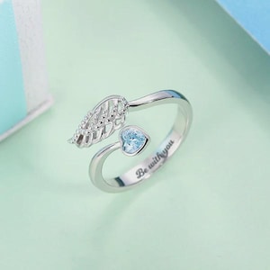 Puede incluir: Anillo plateado con dise&ntilde;o abierto. El anillo presenta un detalle de ala y una piedra preciosa en forma de coraz&oacute;n azul claro. Las palabras "Be with you" est&aacute;n grabadas en la banda. El anillo est&aacute; sobre un fondo azul claro.