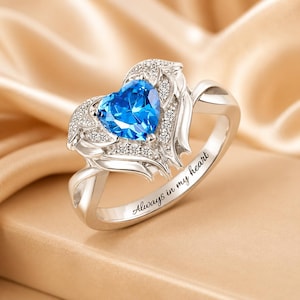 May include: A silver ring featuring a heart-shaped, bright blue gemstone. The heart is surrounded by smaller, clear stones and wing-like designs. The inscription "Always in my heart" is engraved on the band.
