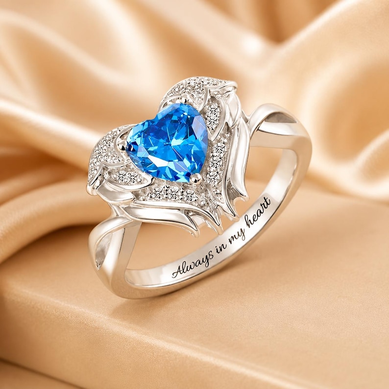 May include: A silver ring featuring a heart-shaped, bright blue gemstone. The heart is surrounded by smaller, clear stones and wing-like designs. The inscription "Always in my heart" is engraved on the band.