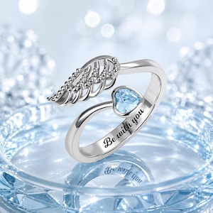 Puede incluir: Un anillo plateado con una gema en forma de coraz&oacute;n azul claro y un dise&ntilde;o de ala de &aacute;ngel. El anillo est&aacute; grabado con las palabras "Be with you". El fondo es borroso y azul claro.