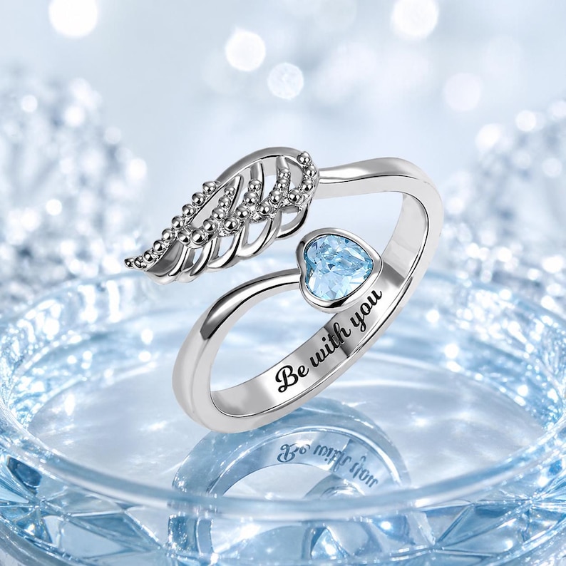 Puede incluir: Un anillo plateado con una gema en forma de coraz&oacute;n azul claro y un dise&ntilde;o de ala de &aacute;ngel. El anillo est&aacute; grabado con las palabras "Be with you". El fondo es borroso y azul claro.