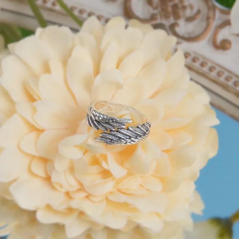 Puede incluir: Un anillo plateado con dise&ntilde;o de plumas. El anillo es abierto y se envuelve alrededor del dedo, con patrones detallados de plumas en cada lado. Se encuentra sobre una flor amarilla crema suave.