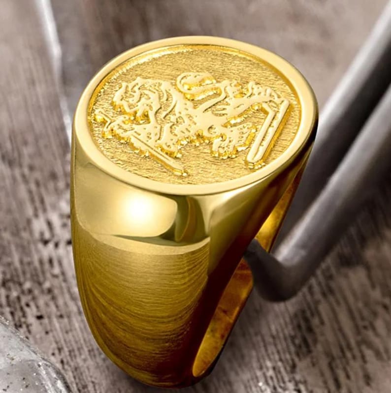 May include: A gold signet ring featuring a circular face with a textured background and a raised, detailed relief of a lion figure. The band of the ring is smooth and polished, showcasing a bright metallic finish throughout the entire piece.
