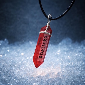 May include: A red crystal pendant with silver wire wrapping, hanging from a black cord. The crystal has etched text and a pointed base. The background is a blurred, icy blue.