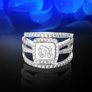 May include: A silver ring with a square-shaped center stone surrounded by smaller stones. The center stone has a monogram engraved on it.