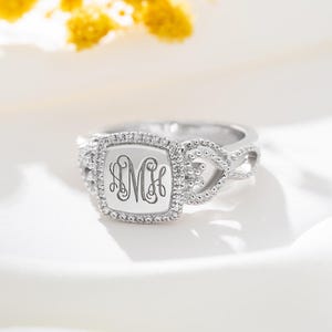 Può includere: Un anello d'argento con una faccia quadrata con un monogramma. La faccia è circondata da piccole pietre trasparenti. La fascia ha un design intrecciato con piccoli accenti di perline. L'anello è posto su uno sfondo bianco morbido.