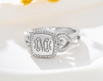 May include: A silver ring with a square face bearing a monogram. The face is encircled by small, clear stones. The band has a braided design with small beaded accents. The ring is set against a soft, white background.
