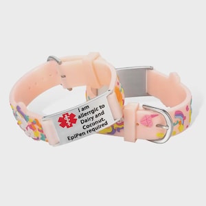 Könnte beinhalten: Zwei hellrosa medizinische Armbänder. Jedes Armband hat eine Metallplatte mit dem roten medizinischen Warnsymbol und dem Text: "I am allergic to Dairy and Coconut. EpiPen required." Die Bänder sind mit bunten Designs verziert.