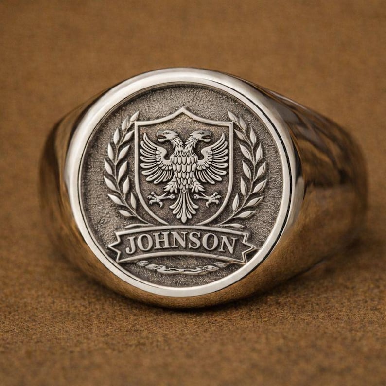 May include: A silver-toned signet ring featuring a circular face with an embossed double-headed eagle emblem inside a shield, flanked by laurel branches. Below the shield, a banner displays the text JOHNSON in raised capital letters.