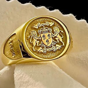 May include: A gold signet ring featuring a circular face with an engraved coat of arms, two lions, a crown, and the text FAMILY KENNEDY. The side of the ring band is engraved with the year 1955 in a bold, recessed font style.