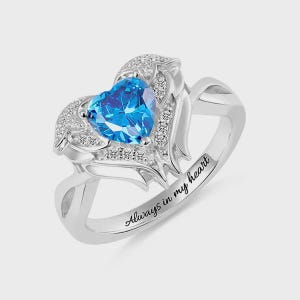 May include: A silver ring featuring a heart-shaped, bright blue gemstone. The gemstone is surrounded by smaller, clear stones and decorative metalwork. The ring band is engraved with the words "Always in my heart."