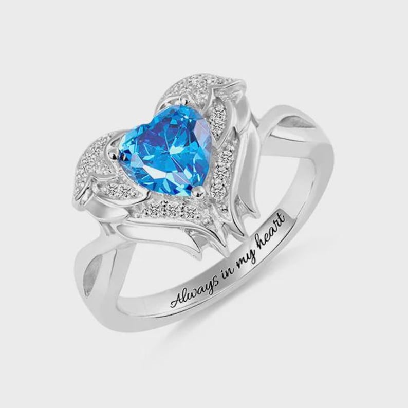 May include: A silver ring featuring a heart-shaped, bright blue gemstone. The gemstone is surrounded by smaller, clear stones and decorative metalwork. The ring band is engraved with the words "Always in my heart."