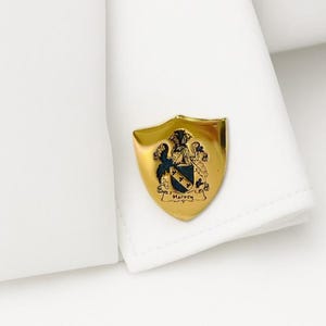 K&ouml;nnte beinhalten: Goldfarbener Manschettenknopf in Schildform mit Familienwappen. Das Wappen zeigt ein Wappen mit einem schwarzen und blauen Schild, zwei gekreuzten Sternen und dem Namen "Harvey". Der Manschettenknopf ist an einer wei&szlig;en Hemdmanschette befestigt.