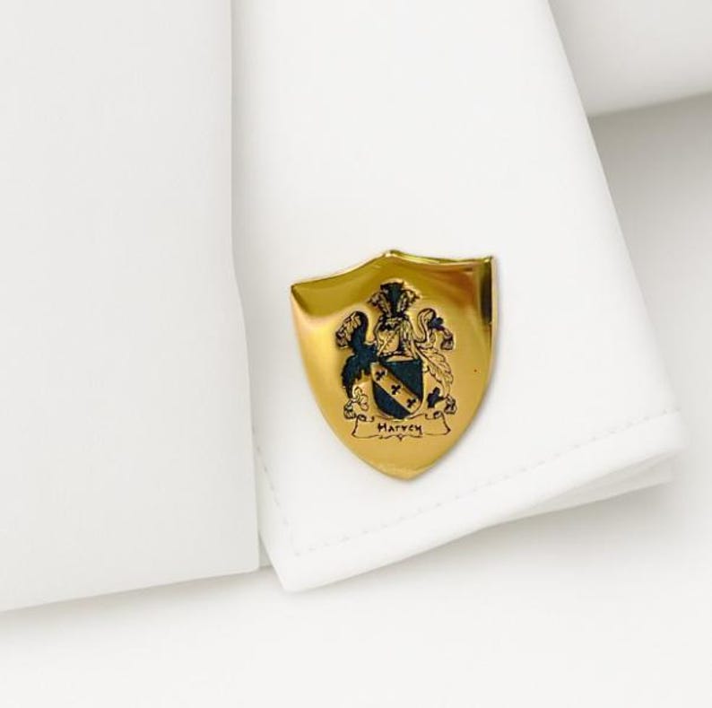 K&ouml;nnte beinhalten: Goldfarbener Manschettenknopf in Schildform mit Familienwappen. Das Wappen zeigt ein Wappen mit einem schwarzen und blauen Schild, zwei gekreuzten Sternen und dem Namen "Harvey". Der Manschettenknopf ist an einer wei&szlig;en Hemdmanschette befestigt.