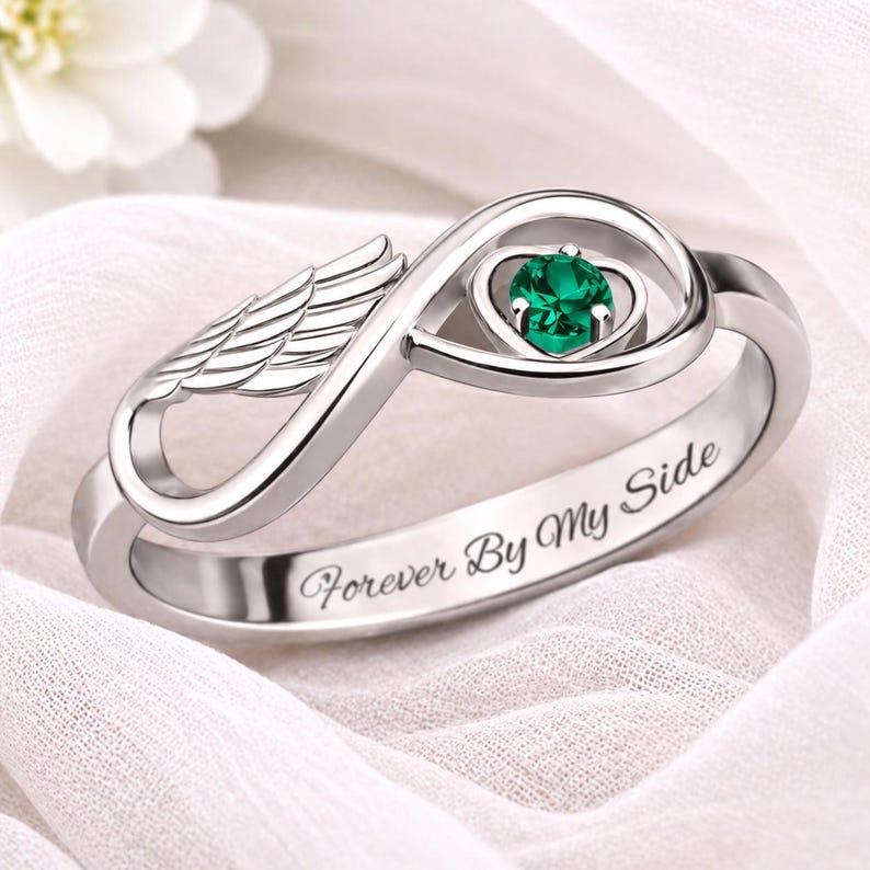 May include: A silver ring with an infinity symbol design, featuring a green gemstone and wing detail. The ring has the inscription "Forever By My Side". The ring is set against a white background.