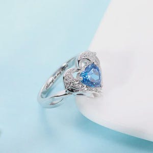 May include: A silver ring with a heart-shaped blue gemstone. The ring features a delicate design with small, clear stones and the inscription "Always in my heart."