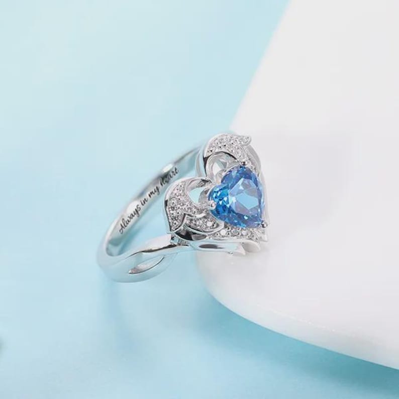 May include: A silver ring with a heart-shaped blue gemstone. The ring features a delicate design with small, clear stones and the inscription "Always in my heart."