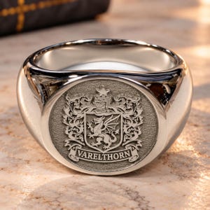 May include: A polished silver-toned signet ring featuring a circular face with an engraved heraldic crest. The design includes a dragon, a knight's helmet, and a star above a banner with the text VARELTHORN in capital letters.