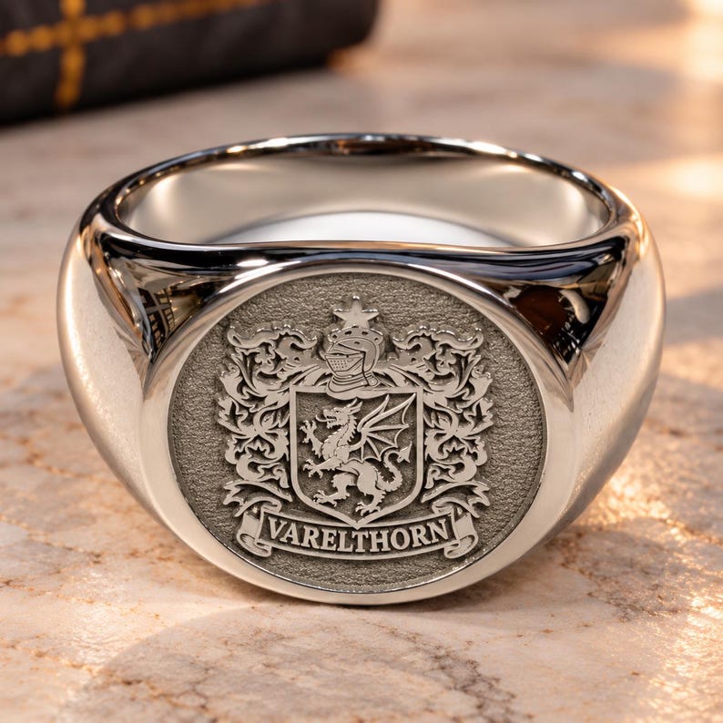 May include: A polished silver-toned signet ring featuring a circular face with an engraved heraldic crest. The design includes a dragon, a knight's helmet, and a star above a banner with the text VARELTHORN in capital letters.