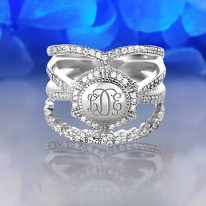 Könnte beinhalten: Ein silberner Ring mit drei Bändern, die jeweils mit kleinen funkelnden Steinen verziert sind. Das mittlere Band hat einen größeren runden Zargen mit einem Monogramm.