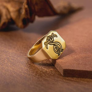 May include: A gold-colored signet ring with an engraved design on the octagonal face. The ring features a detailed, dark-colored emblem. The ring is set against a brown, textured surface, with a blurred background.