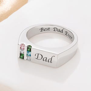 Op de afbeelding: Een zilveren ring met de inscriptie "Best Dad Ever" en "Dad". De ring heeft drie gekleurde edelstenen: roze, groen en blauw. De ring heeft een modern, minimalistisch ontwerp.